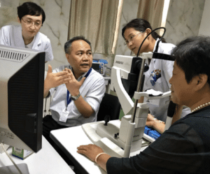 Three people gathered around a man speaking with medical eye equipment on the desk.