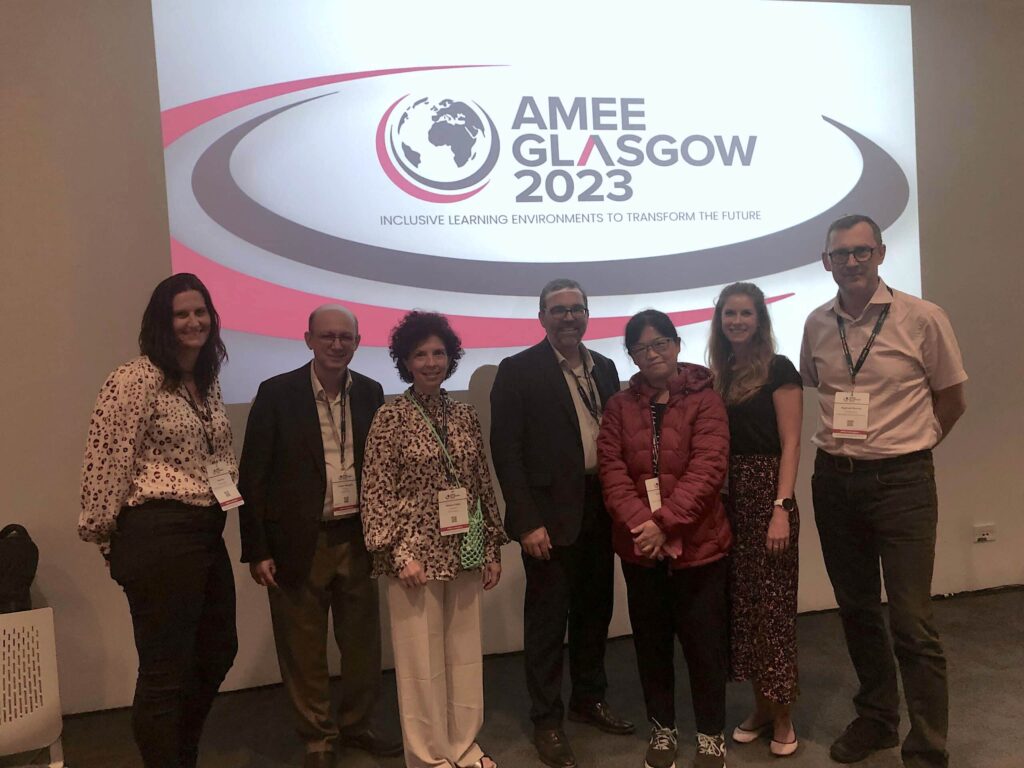 seven people standing on stage with a projection behind them that says AMEE Glasgow 2023